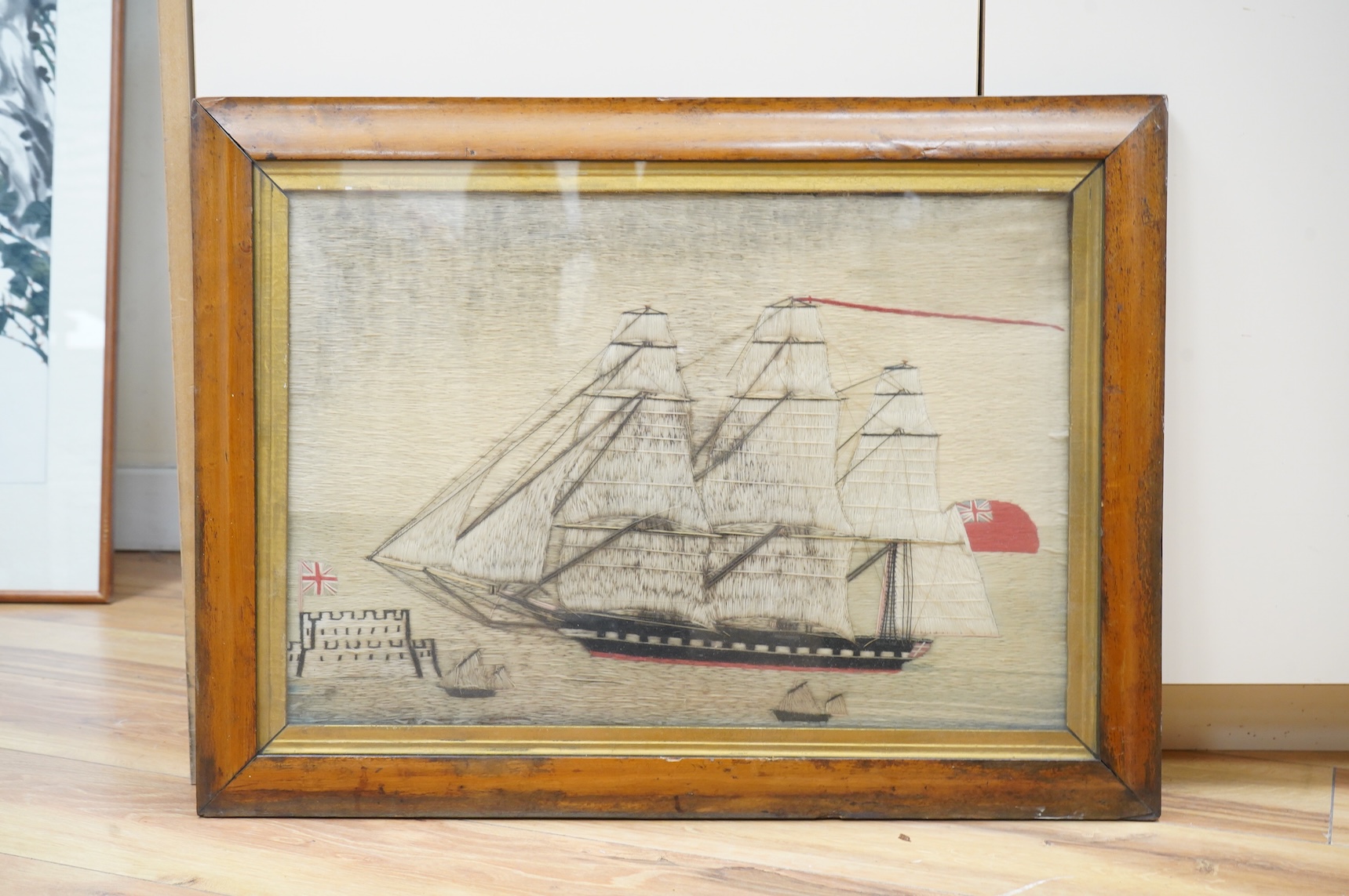 A 19th century maple framed Folk Art, sailors wool work or ‘Woolie’ of a Royal Navy cutter sailing past smaller ships in the foreground and a castle flying a Union Jack, 48cm wide x 33cm high. Condition - possibly hung o